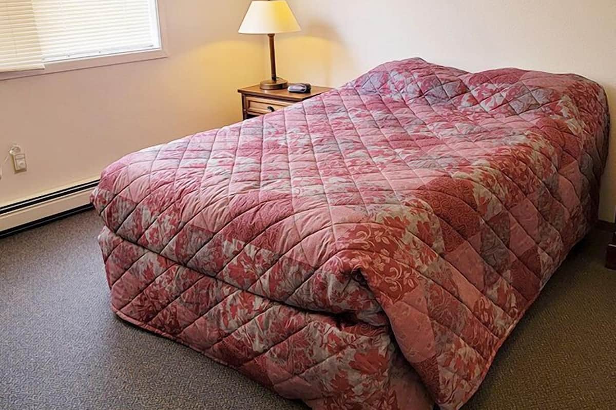 A bedroom with a bed, a nightstand, a lamp, and a window with blinds.