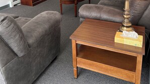 A living room with a brown sofa, a wooden coffee table, a lamp, a TV on a wooden cabinet, and a potted plant. - Adak Island Inn (Adak Island)