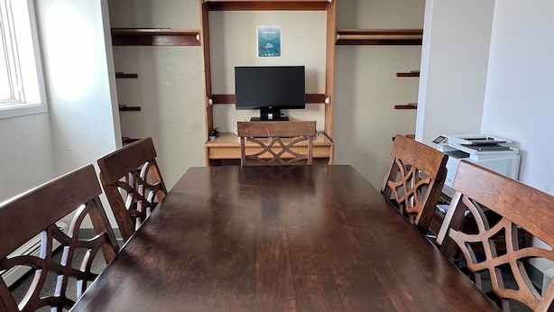 A conference room with a wooden table and chairs, a television on a stand, and a bulletin board with a sign. - Adak Island Inn (Adak Island)