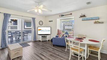 Sala de estar con televisor, una mesa de comedor con sillas, sofá, ventilador de techo y vista al exterior.