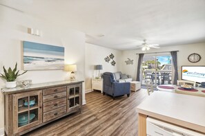 A modern living room with a wooden cabinet, a blue armchair, a dining area with a table and chairs, and a balcony with a view. - Townhome · Steps to Gulf Beach & Pool (Port St. Joe)
