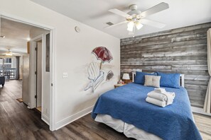 A bedroom with a bed, a ceiling fan, a wooden wall, and a painting of an umbrella. - Townhome · Steps to Gulf Beach & Pool (Port St. Joe)