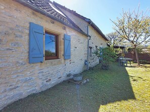 A stone house with a window and shutters, a small garden, and a tree.