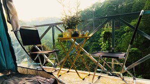 Ein Zelt mit Blick auf einen Wald. Im Inneren befinden sich ein Klappstuhl, ein kleiner Tisch mit einer Pflanze und ein Schlafbereich mit einer Decke.