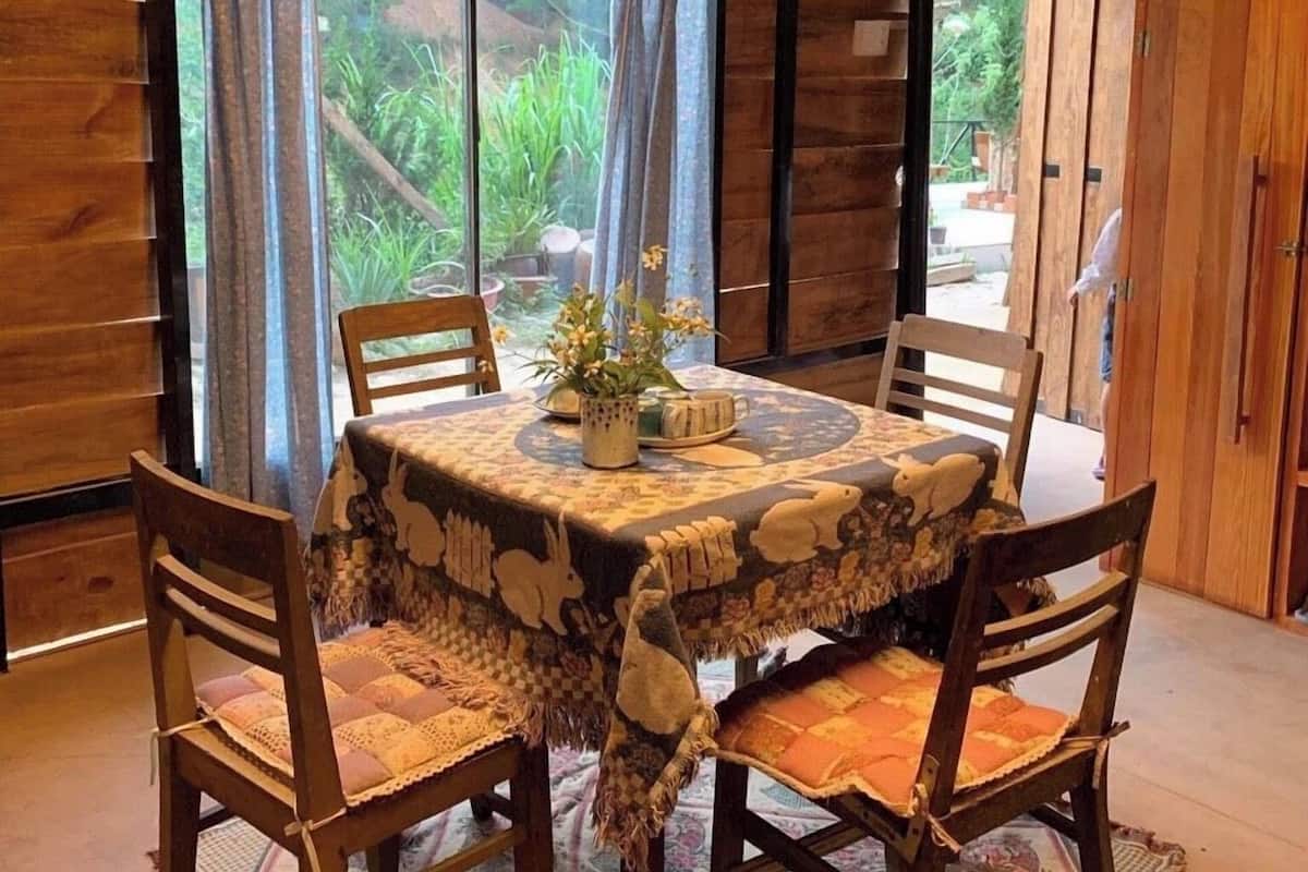 Uma área de refeições com mesa de madeira coberta por toalha estampada, rodeada por quatro cadeiras de madeira.