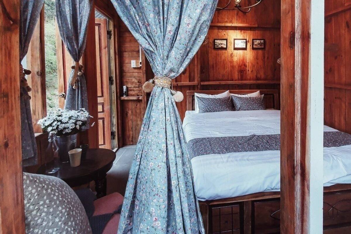 Quarto de cabana de madeira com cama, cortinas, uma pequena mesa com um vaso e uma cadeira.