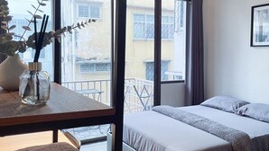 A bedroom with a bed, a bedside table with a vase, and a window with a view of buildings. - MAXi MaMa Room (Bangkok)