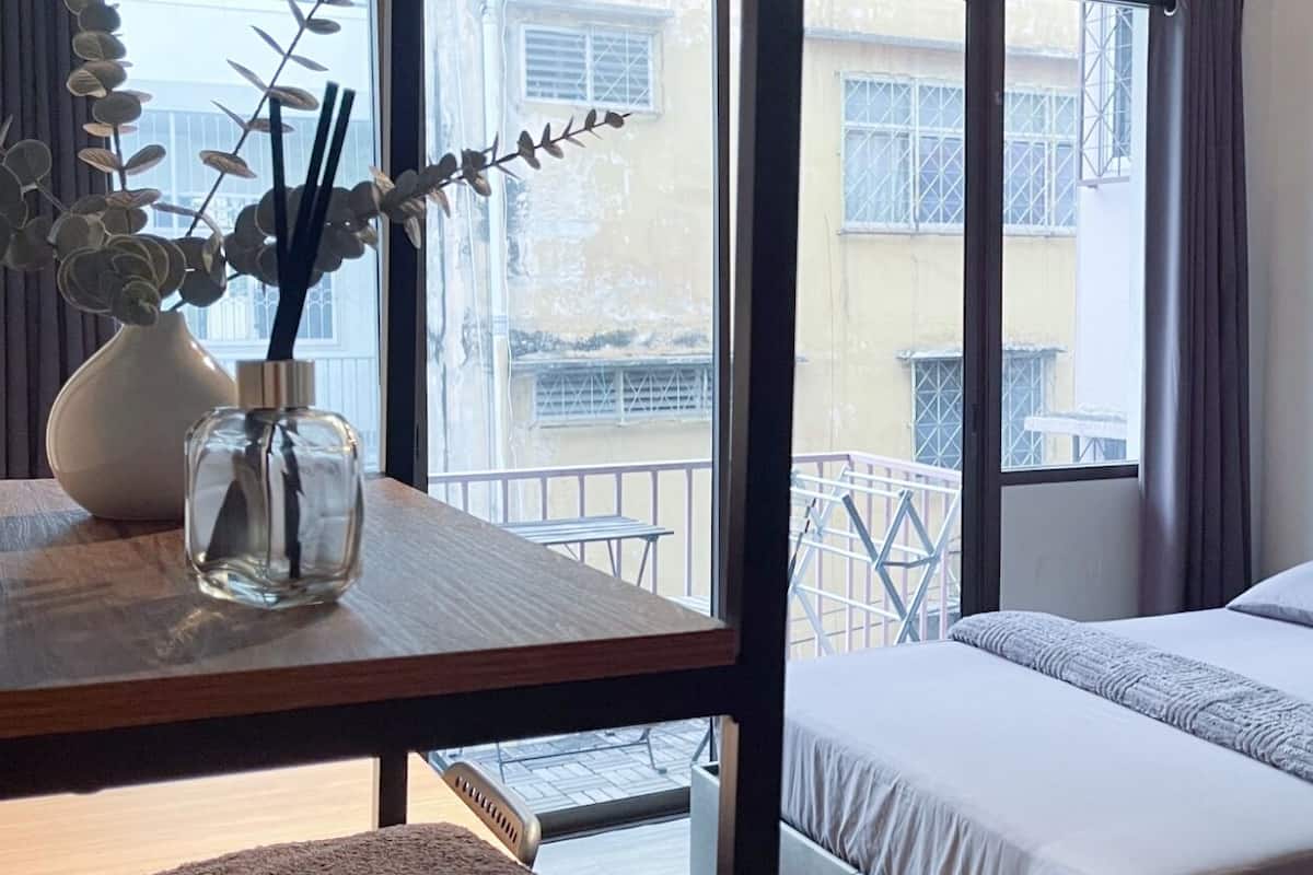 Une chambre à coucher avec un lit, une table de chevet ornée d’un vase et une fenêtre donnant sur des immeubles.