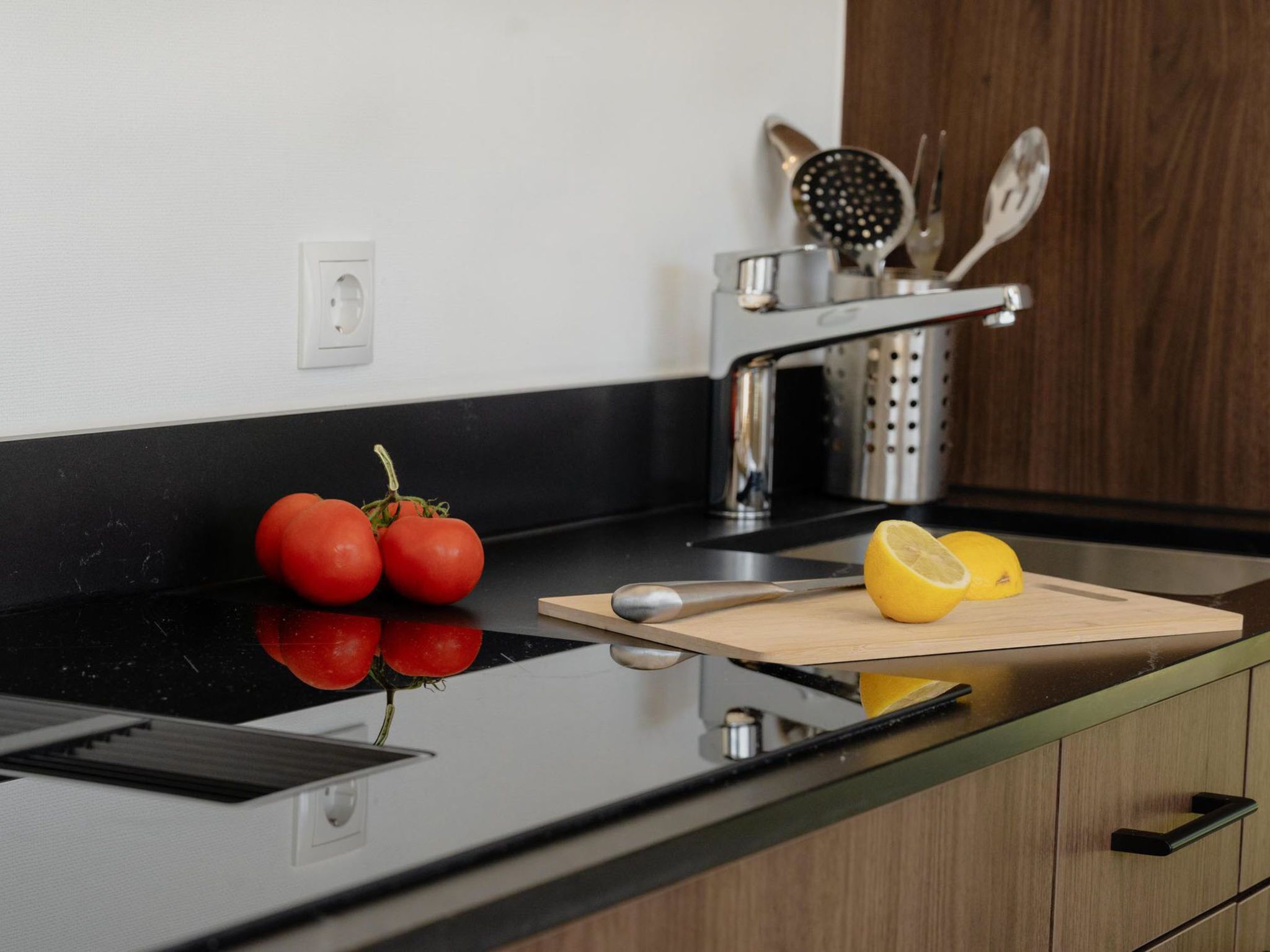 Une cuisine avec un plan de travail noir, une planche à découper, un citron et des tomates.