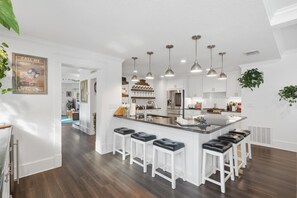 A modern kitchen with white cabinetry, a central island with bar stools, pendant lighting, and a visible refrigerator.