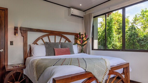 Une chambre avec un lit en bois, une table de chevet, une commode, une fenêtre donnant sur un paysage verdoyant et un appareil de climatisation.