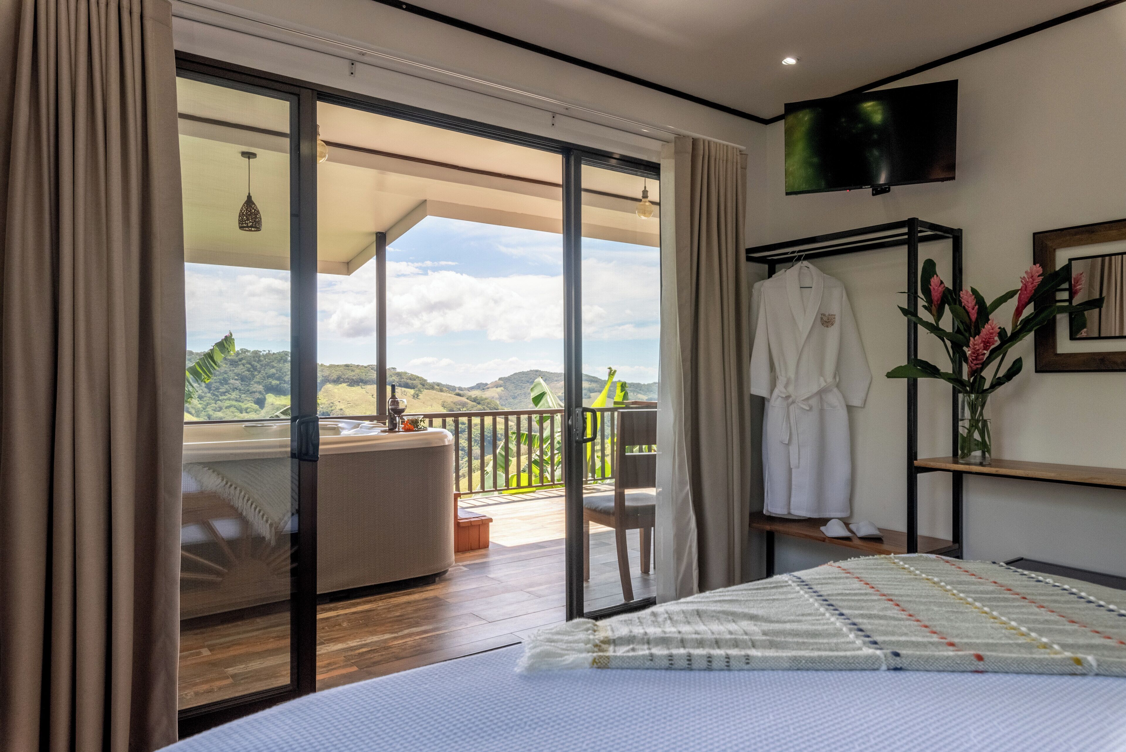 Un dormitorio con una cama grande, un balcón con vistas, un televisor instalado en la pared y una bata colgada en un perchero.