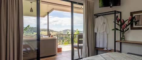 Un dormitorio con una cama grande, un balcón con vistas, un televisor instalado en la pared y una bata colgada en un perchero.