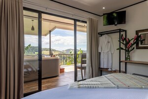 Un dormitorio con una cama grande, un balcón con vistas, un televisor instalado en la pared y una bata colgada en un perchero.