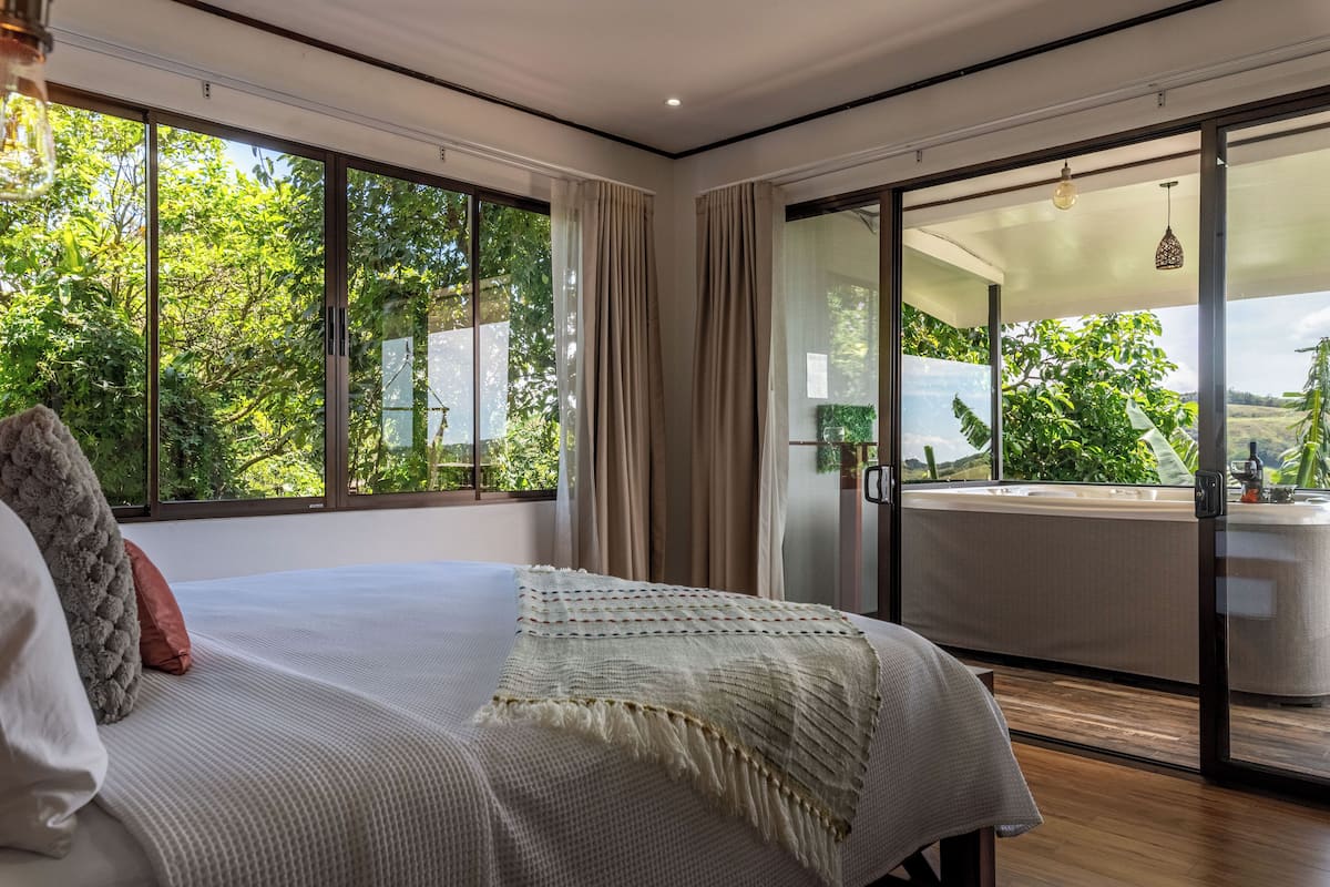 Une chambre moderne avec un grand lit, un balcon doté d’un jacuzzi et une vue sur la verdure.