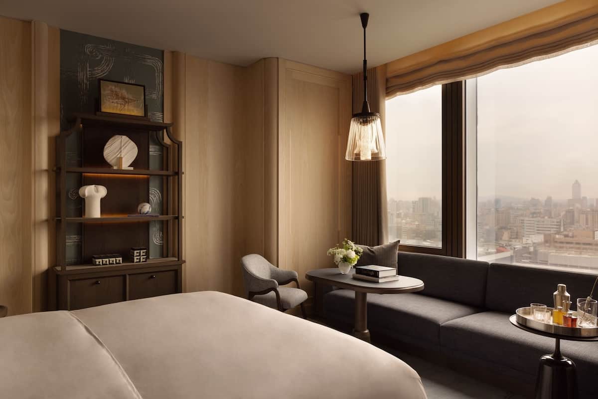 A modern hotel room with a large bed, a sofa, a small table, and a bookshelf. There is a city view through the window.