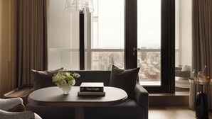 Quarto de hotel moderno com uma grande janela que oferece vista da cidade, uma mesa redonda com um livro e flores, e uma luminária pendente elegante.