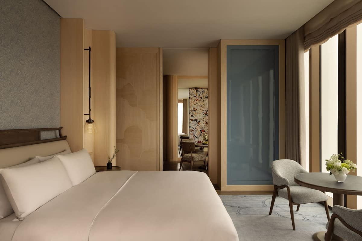 A hotel room with a large bed, a sitting area with a chair and table, and a view through an open door.