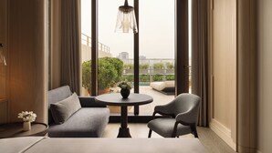 Quarto de hotel moderno com uma grande janela, um sofá, duas cadeiras, uma mesinha redonda e um candeeiro suspenso.