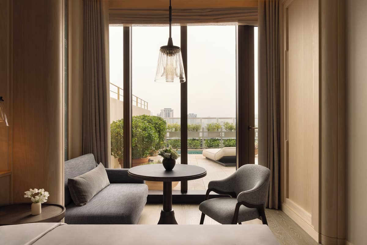 A modern hotel room with a large window, a sofa, two chairs, a small round table, and a hanging light fixture.