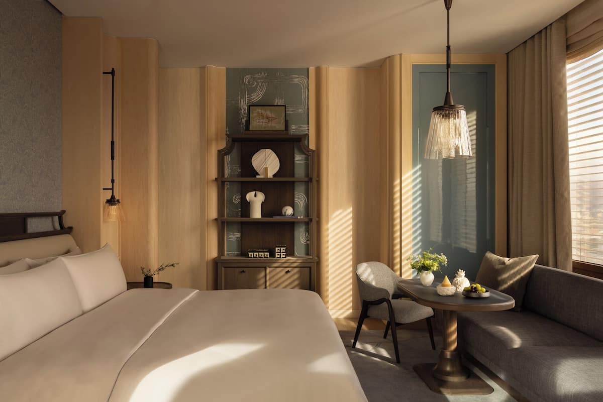 A hotel room with a large bed, a seating area with a table, a decorative shelf, and a hanging light fixture.
