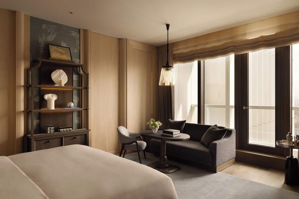 A modern hotel room with a large bed, a seating area with a sofa and armchair, a small table, and a bookshelf.