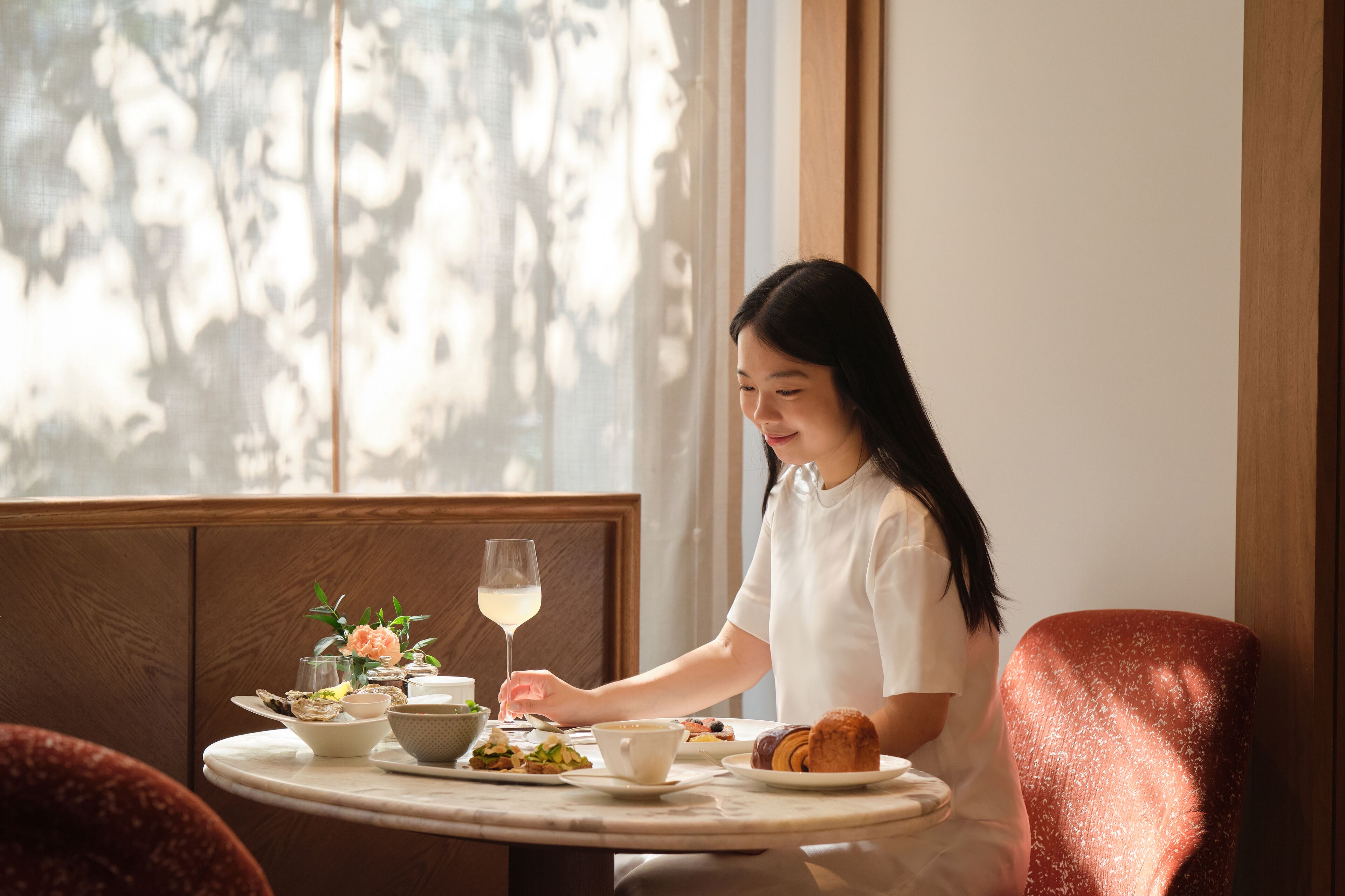 A person seated at a round table with a meal, a glass of white wine, and a flower arrangement.