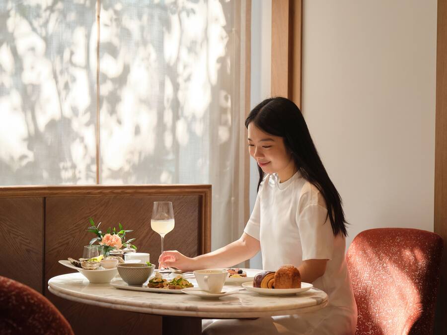 A person seated at a round table with a meal, a glass of white wine, and a flower arrangement.