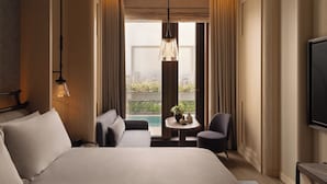 Quarto de hotel com uma cama grande, área de estar com sofá e poltronas, pequena mesa com objetos decorativos, televisão fixada na parede e vista para uma piscina pela janela.
