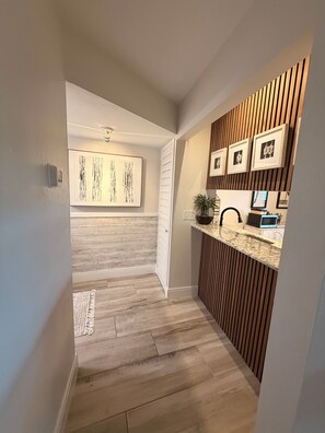 A hallway with a wooden floor, a sink area with a countertop, and a wall-mounted light fixture.
