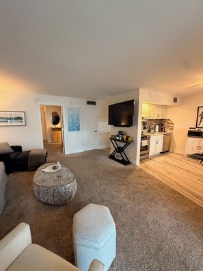 Ein Wohnzimmer mit einer Couch, einem Hocker und einem Fernseher. Es gibt eine Küchenzeile mit Schränken und einer Mikrowelle.