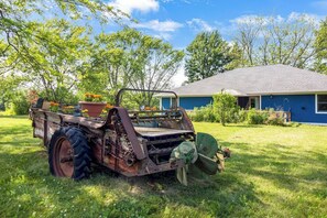 Un tractor antiguo con un remolque en una zona cubierta de hierba, una casa azul al fondo.