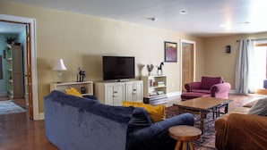 A living room with a blue sofa, a wooden coffee table, a television, and a pink chair. - Spacious 4BR Fennville Farmhouse w/ Pool Table (Fennville)