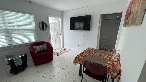 Una sala de estar con un sofá rojo, una mesa de centro de vidrio, un televisor montado en la pared y una zona de comedor con mantel floral.