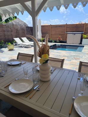 An outdoor dining area with a table set for two, a pool in the background, and a wooden fence enclosing the space.