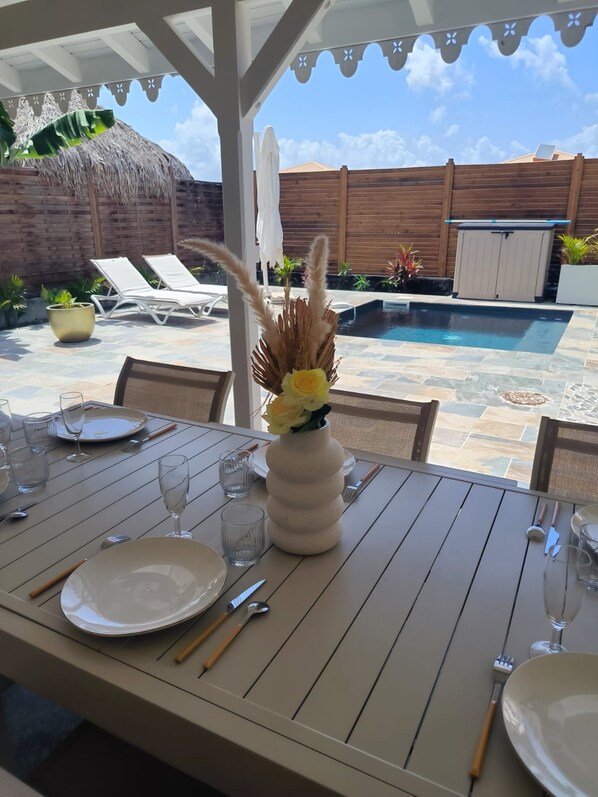 An outdoor dining area with a table set for two, a pool in the background, and a wooden fence enclosing the space.