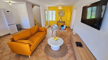 Un salon comprenant un canapé, des fauteuils, une table basse avec un panier de fruits, un téléviseur et un escalier menant à une autre pièce.