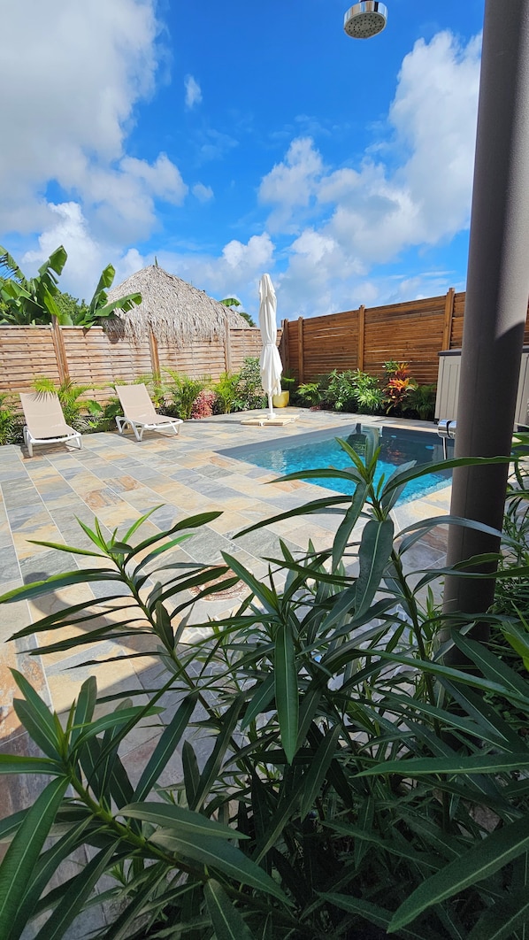 A swimming pool with lounge chairs, a thatched-roof structure, and tropical plants.
