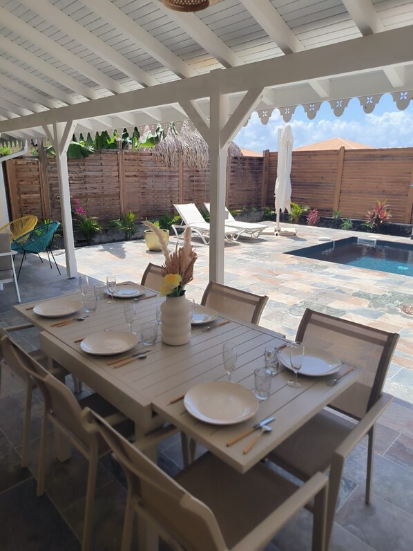 An outdoor patio with a dining table set for four, surrounded by chairs, a pool, and lounge chairs.