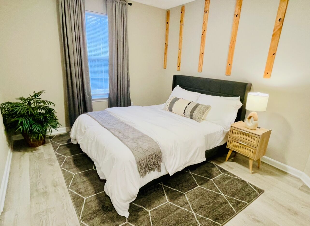 A bedroom with a bed, a nightstand, a lamp, a potted plant, and a window with curtains.