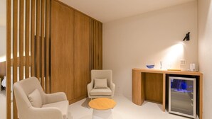 Habitación de hotel moderna con un separador de madera, dos sillones, una mesita redonda, un minibar y un televisor montado en la pared.