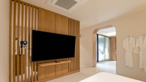 Una habitación de hotel moderna con un televisor de pantalla plana grande montado en una pared de paneles de madera, una mesita de noche con un control remoto y un vestidor con batas colgadas.