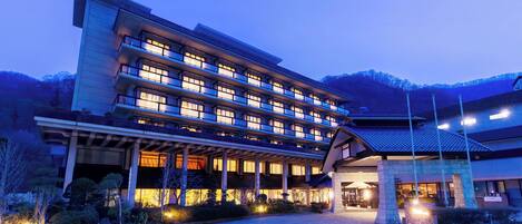 A multi-story hotel with a traditional Japanese architectural style, featuring a large entrance with a covered walkway and a stone building adjacent to it.