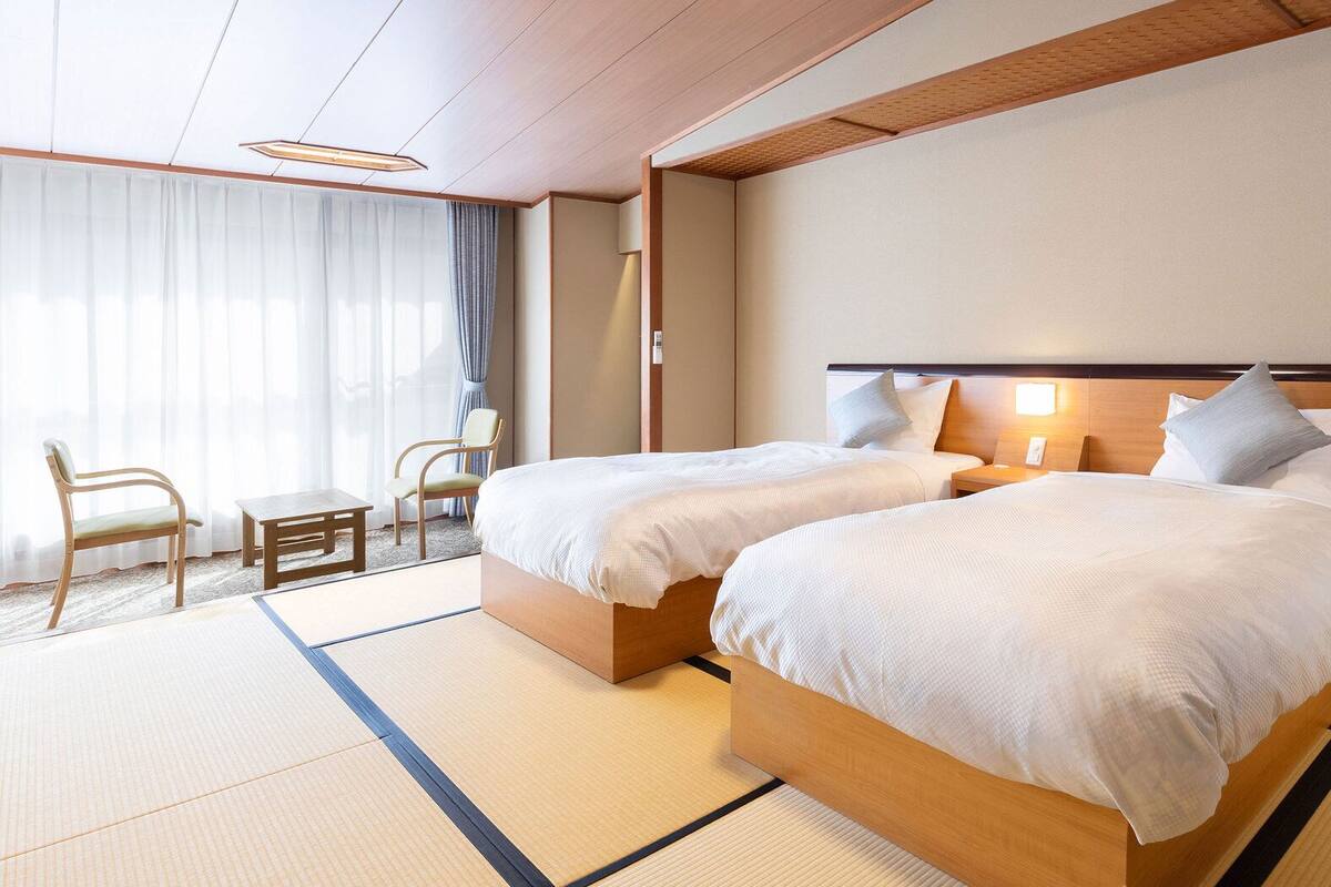 A hotel room with two beds, a small table, chairs, and a view of the outdoors.