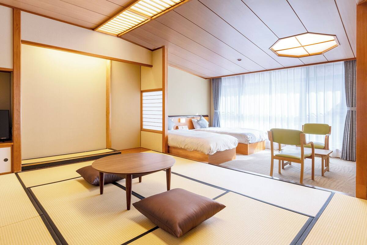 A traditional Japanese-style hotel room with tatami flooring, a bed, a television, and a view of the city.