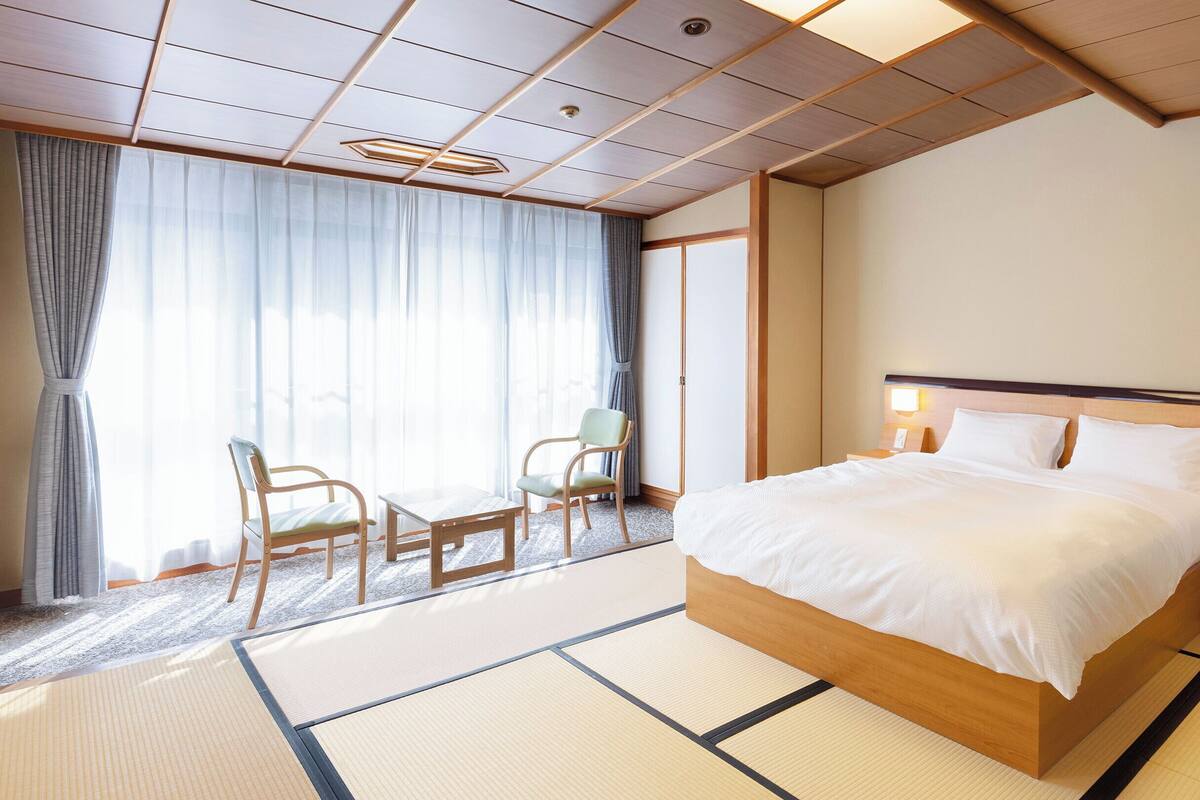A hotel room with a large bed, a seating area with chairs, a view of the outdoors, and a traditional Japanese-style ceiling.