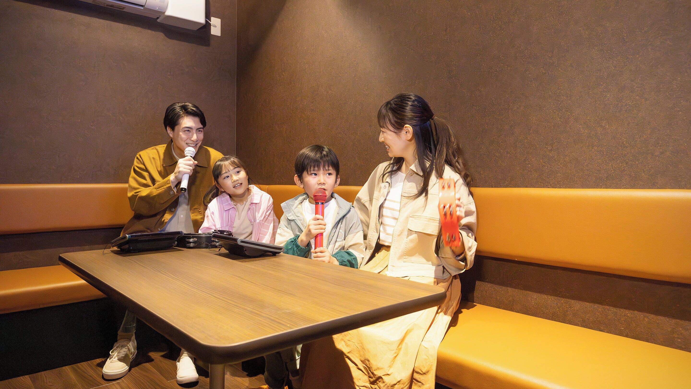 A group of people seated at a wooden table in a room with brown walls and yellow cushioned seating.