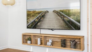 Un televisor de pantalla plana montado en una pared blanca, mostrando una escena de playa con un paseo marítimo de madera. Debajo del televisor, hay un estante de madera con objetos decorativos.