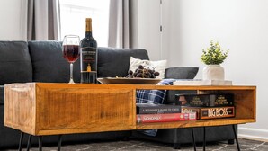 Una mesa de centro de madera con una botella de vino y una copa, un cuenco de uvas y juegos de mesa frente a un sofá gris.