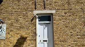 A brick building with a white door and window, a small sign on the wall, and a shadow on the ground. - Eurostar Private Flat Sleeps up to 4 London (London)